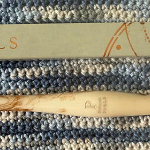May include: A cream-colored crochet hook with brown marbling, next to a light green box with the word "FURLS" in gold lettering. The hook is on a blue and white crocheted surface.