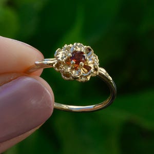 Pode incluir: Um anel dourado com um design floral e uma pedra vermelha central. A banda do anel é fina e a configuração floral tem detalhes intrincados. O anel é exibido em um fundo verde desfocado.