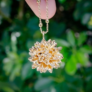 May include: A gold-colored pendant necklace featuring a detailed flower design. The pendant is a three-dimensional rendering of a flower with multiple layers of petals. The necklace chain is delicate, with small, round beads interspersed along its length.