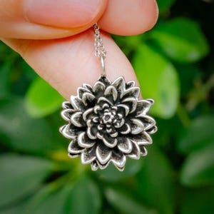 May include: A silver-colored pendant necklace featuring a detailed, three-dimensional flower design. The flower has multiple layers of petals, creating a textured appearance. The necklace is held by a delicate chain, and the background is blurred green foliage.