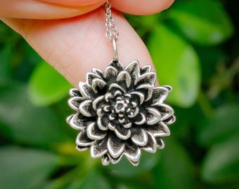 Pode incluir: Um colar com pingente prateado com um design floral detalhado. A flor tem múltiplas camadas de pétalas, criando uma aparência texturizada. O colar é preso por uma corrente delicada. O fundo é uma folhagem verde desfocada.