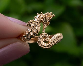 May include: A gold-colored seahorse ring. The ring features a detailed seahorse design with a textured surface. The seahorse is curled around the finger, with the head and tail meeting. The ring is set against a blurred green background.