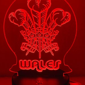 Puede incluir: Luz nocturna LED roja con el dragón galés, un dragón rojo con una corona y el texto "WALES" debajo. La luz está sobre una base negra con un cable.