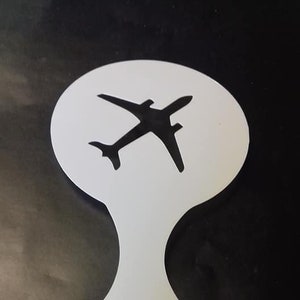 Puede incluir: Plantilla de plástico blanco con una silueta de un avión. La plantilla tiene un mango con un agujero en el centro.