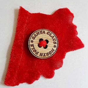 Puede incluir: Un botón de madera con el texto "Santa Claus North Pole" y un hilo rojo en el centro. El botón está sobre un fondo de tela roja.