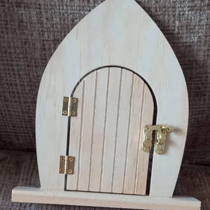 May include: A wooden fairy door with a rounded top and a small, arched door. The door has vertical wooden slats and is secured with brass hinges and a latch. The frame is light-colored wood, and the door is designed to be a decorative item.
