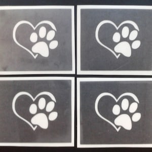 Op de afbeelding: Vier witte vinyl sjablonen met een pootprint in een hartvormig ontwerp. De sjablonen zijn op een grijze achtergrond.
