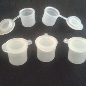 Könnte beinhalten: Acht kleine, transparente Plastik-Farbtöpfe mit Deckeln. Die Deckel haben einen kleinen Griff an der Seite.