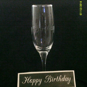 Può includere: Un flauto da champagne in vetro trasparente con le parole "Happy Birthday" incise sul lato. Il flauto è appoggiato su una superficie nera con un cartello nero e bianco che recita "Happy Birthday".