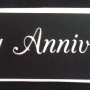 Puede incluir: Un letrero rectangular negro con las palabras "Happy Anniversary" en elegante escritura blanca. El letrero tiene un borde blanco, creando un diseño limpio y clásico, adecuado para celebrar una ocasión especial.