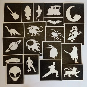 Op de afbeelding: Een set van 20 zwart-wit sjablonen met verschillende ontwerpen, waaronder een gitaar, een draak, een spin, een trein, een dinosaurus, een planeet, een auto, een voetballer, een leeuw, een alien, een hagedis, een geest, een vleermuis, een halve maan, een vlam, een krijger, een piraat en een karateka.