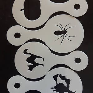 May include: A collection of four white plastic stencils with Halloween-themed cutouts. Designs include a pumpkin, spider, bat, and witch on a broom. Each stencil has a handle and a small hole for hanging. The stencils are arranged on a black surface.