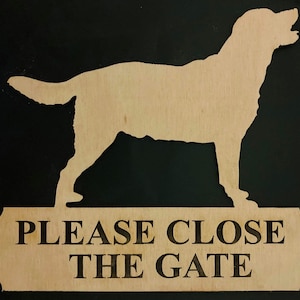 Op de afbeelding: Houten bord met een silhouet van een hond en een plaatje eronder. Op het plaatje staat "PLEASE CLOSE THE GATE" in het zwart. Het bord is gemaakt van lichtgekleurd hout, tegen een donkere achtergrond.