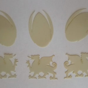 Op de afbeelding: Drie witte, uitgesneden rugbyballen van plastic en drie witte, uitgesneden Welshe draken van plastic.