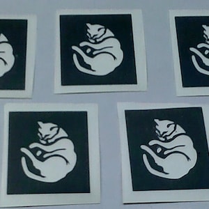 Puede incluir: Cinco pegatinas cuadradas con un gato blanco enroscado sobre un fondo negro. Cada pegatina tiene un borde blanco. Las pegatinas están dispuestas sobre una superficie clara. El diseño del gato es simple y elegante.