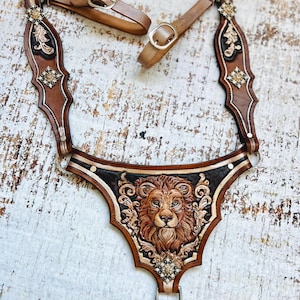 May include: A brown leather halter with a lion head design and gold accents. The halter has a decorative strap with a buckle and a separate strap with a ring.