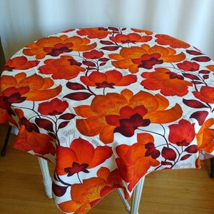 May include: A white tablecloth with a large orange and brown floral pattern. The tablecloth is draped over a round table with white legs.