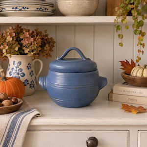 May include: A matte blue ceramic lidded pot with a rounded body, two side handles, and a central arched handle on the lid. The pot features horizontal embossed lines around its circumference and sits on a white surface with a rustic aesthetic.