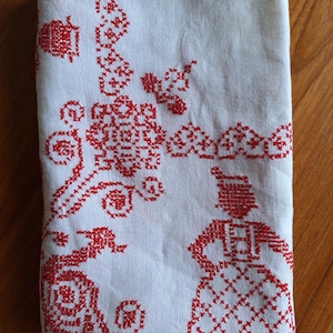 May include: Folded white linen cloth with intricate red cross-stitch embroidery. The design features floral and geometric patterns, along with a stylized figure. The cloth is folded, showing the detailed needlework.