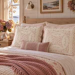 May include: Two rectangular quilted pillow shams featuring a cream-coloured centre with a small floral print, surrounded by a scalloped pink border. A long, rectangular, textured dusty rose lumbar pillow sits in front of the two larger shams.