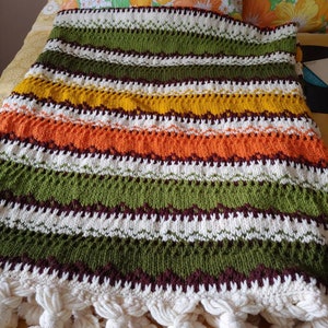 May include: A striped knitted throw blanket with a fringe border in shades of green, orange, yellow, white, and brown.