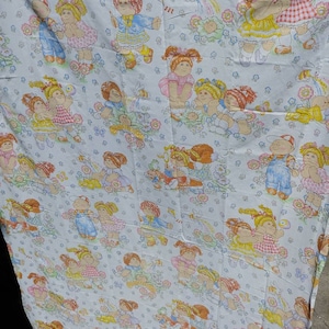 Pode incluir: Um lençol branco de tamanho individual com um padrão repetido de bonecas Cabbage Patch Kids em várias poses e roupas. As bonecas são cercadas por flores, borboletas e outros detalhes caprichosos.