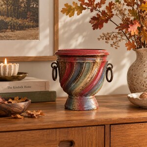 May include: A ceramic urn-shaped planter featuring a multi-coloured, swirled glaze in shades of red, blue, and gold. The vessel has a wide, flared rim, a pedestal base, and two dark metal ring handles attached to the sides of the body.