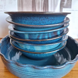 May include: A stack of four deep blue ceramic bowls with a speckled finish and dark brown rims, resting inside a larger, wide, shallow blue ceramic bowl with a scalloped edge and a ribbed texture on the exterior surface.
