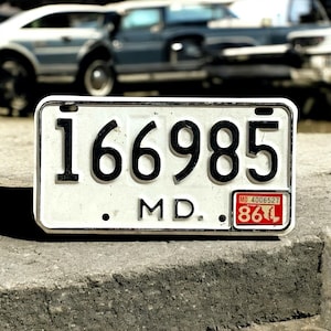 May include: A rectangular white license plate with black numbers and letters. The plate reads "166985" above "MD." and has a red and white sticker in the bottom right corner.