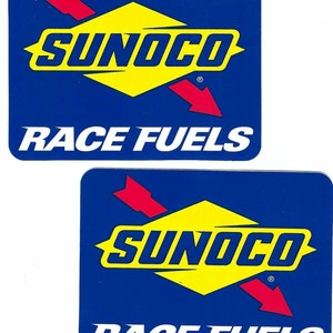 May include: Two blue and yellow Sunoco Race Fuels stickers. Each sticker features a yellow diamond with the word Sunoco in white letters and a red arrow pointing down. The text "Race Fuels" is in white letters below the diamond.
