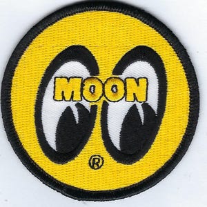May include: A circular embroidered patch featuring a bright yellow background with a black border. In the center, two stylized white and black eye shapes are depicted with the yellow text MOON overlaid. A small registered trademark symbol is below.