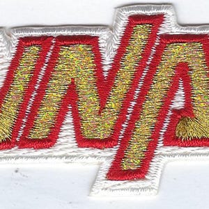 May include: A rectangular embroidered patch featuring the word LUNATI in gold thread with a bold red outline. The letters are set against a white background with a thick white border, creating a clean and distinct look for this fabric accessory.
