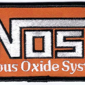 May include: A rectangular embroidered patch with an orange background and a black border. The patch features the white text NOS with an arrow extending from the S, and the white text Nitrous Oxide Systems printed underneath in a serif font.