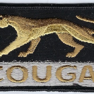 May include: A rectangular embroidered patch featuring a gold-colored cougar in a prowling stance on a black background. Below the cougar, the word COUGAR is stitched in gold thread against a light gray horizontal bar. The entire patch has a black border.