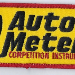 May include: A rectangular embroidered patch with a yellow background and a red border. It features the black text Auto Meter and the red text COMPETITION INSTRUMENTS, along with a black and white graphic icon of a gauge on the left side.