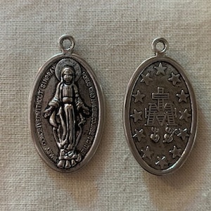 May include: Two silver metal medallions with religious imagery. One features a depiction of the Virgin Mary with the text "Omne Datum Optimum a Deo Patre per Jesum Christum" around the edge. The other features a stylized image of the Virgin Mary with a star pattern around the edge.