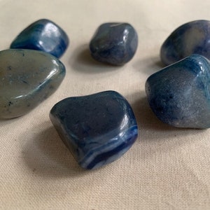 May include: Six smooth, polished blue agate stones with white and gray banding. The stones are various shapes and sizes, and are arranged in a circle on a white fabric background.