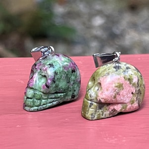 May include: Two green and pink skull pendants with silver bail. The skulls are carved from a green and pink stone with a speckled pattern.