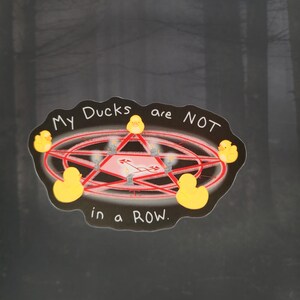 May include: A sticker with the text "My Ducks are NOT in a ROW." The design features a red pentagram with yellow rubber ducks at each point. The background is a blurred image of a forest.