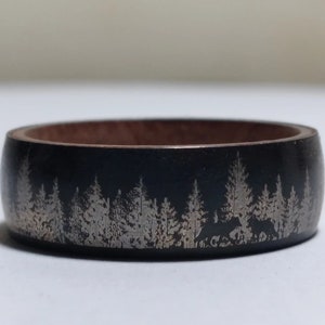 May include: A black ring with a silver design of a forest with trees and a bear. The ring is made of wood and metal.