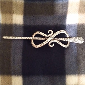 May include: A silver-toned metal hair pin with a decorative, swirling design. The pin is textured with small dots.