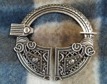 May include: A silver-toned circular brooch with intricate Celtic knotwork designs. The brooch is made of metal and has a circular shape with two curved arms.