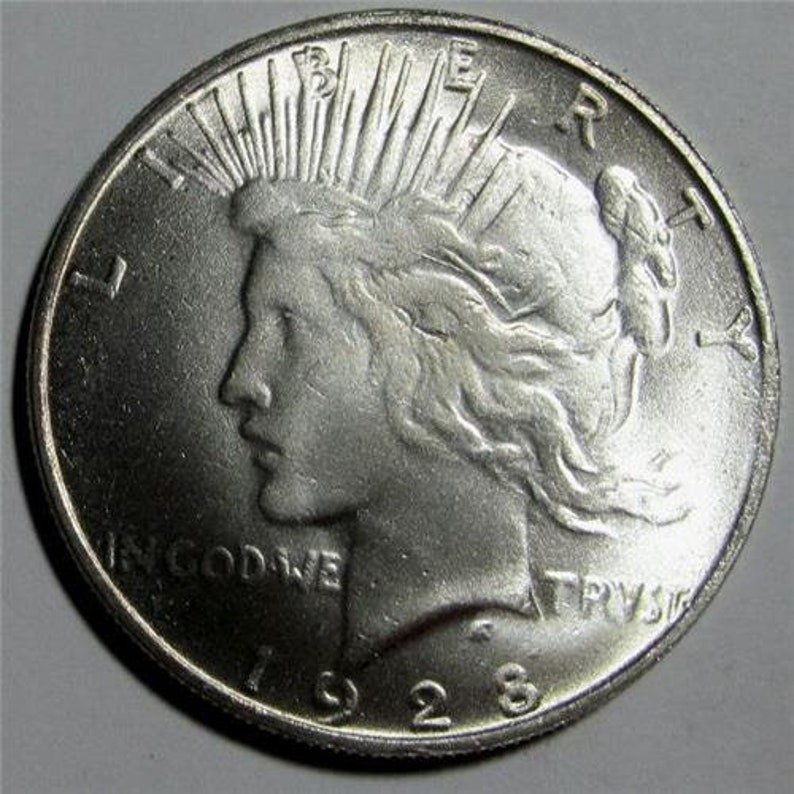 May include: A silver Peace dollar coin with the year 1928 inscribed on the bottom. The coin features a profile of a woman with flowing hair and the words "In God We Trust" and "Liberty" inscribed on the coin.