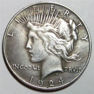 May include: A silver Peace dollar coin with the year 1924 inscribed on the bottom. The coin features a profile of a woman with flowing hair and a crown of rays. The inscription "LIBERTY" is on the right side of the coin and "IN GOD WE TRUST" is on the left side.