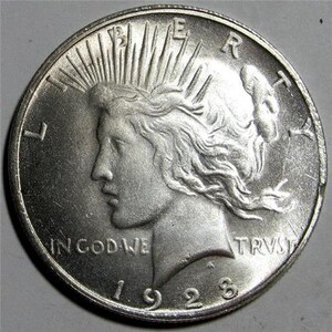 May include: Silver Peace Dollar coin with a profile of Liberty on the obverse. The coin has a radiating sunburst design and the inscription "LIBERTY" on the right side. The inscription "IN GOD WE TRUST" is at the bottom of the coin. The date "1928" is at the bottom center of the coin.