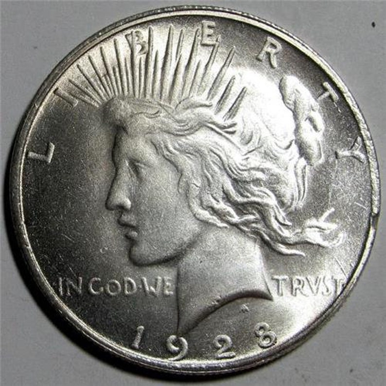 May include: Silver Peace Dollar coin with a profile of Liberty on the obverse. The coin has a radiating sunburst design and the inscription "LIBERTY" on the right side. The inscription "IN GOD WE TRUST" is at the bottom of the coin. The date "1928" is at the bottom center of the coin.