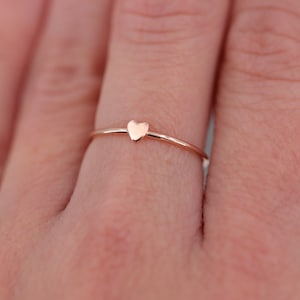 Può includere: Un delicato anello in oro rosa con un piccolo design a forma di cuore. L'anello è indossato su un dito.