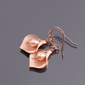 K&ouml;nnte beinhalten: Ein Paar ohrringe in ros&eacute;goldfarbener Optik, geformt wie Calla-Lilien-Bl&uuml;ten, jeweils mit einer zentralen wei&szlig;en Perle. Das Metall hat eine geb&uuml;rstete, matte Oberfl&auml;che und ist an geschwungenen Ohrhaken befestigt, was ein florales Schmuckst&uuml;ck ergibt.