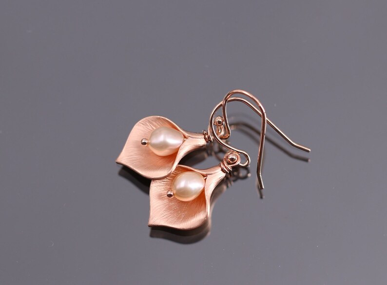 K&ouml;nnte beinhalten: Ein Paar ohrringe in ros&eacute;goldfarbener Optik, geformt wie Calla-Lilien-Bl&uuml;ten, jeweils mit einer zentralen wei&szlig;en Perle. Das Metall hat eine geb&uuml;rstete, matte Oberfl&auml;che und ist an geschwungenen Ohrhaken befestigt, was ein florales Schmuckst&uuml;ck ergibt.