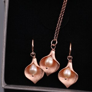 K&ouml;nnte beinhalten: Ein Schmuckset bestehend aus einer Halskette und zwei Ohrh&auml;ngern in Ros&eacute;gold-Optik. Jedes St&uuml;ck zeigt ein strukturiertes, mattes Calla-Lilien-Bl&uuml;tendesign mit einer einzelnen, glatten, cremefarbenen Perle, die in der Mitte des Bl&uuml;tenblatts sitzt.